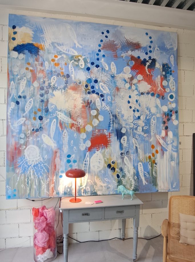 Pintura abstracta con tonos azules, rojos y detalles en blanco, acompañada de una lámpara y mesa.